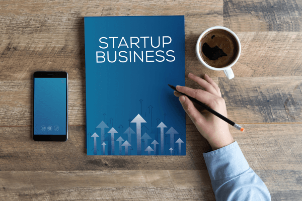 Caderno escrito Startup Business. Caneca de um lado e celular do outro.